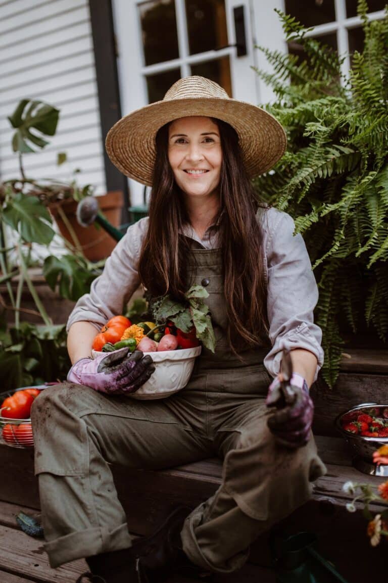 Woman in straw hat and overalls with basket of vegetables in a garden.